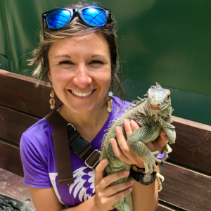 Photo of Jackie Childers, the author. Smiling woman in purple shirt and sunglasses holding an iguana
