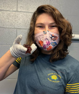 Photo of Lauren E. Simonitis holding a sea hare in a gloved hand. Lauren has brown curly shoulder-length hair and is wearing a mask with a fish pattern on it.