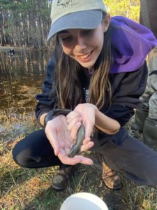 Megan crouching next to a pond, displaying a tadpole in her cupped hands.