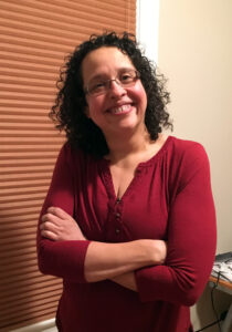 Patricia Hernandez is a Latinx woman wearing a red shirt, smiling while standing next to a window